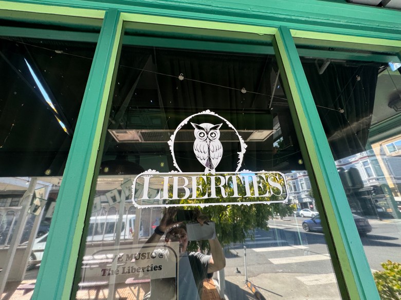 A window decal for “Liberties” featuring an owl logo above the text, with a person holding a phone visible in the reflection.