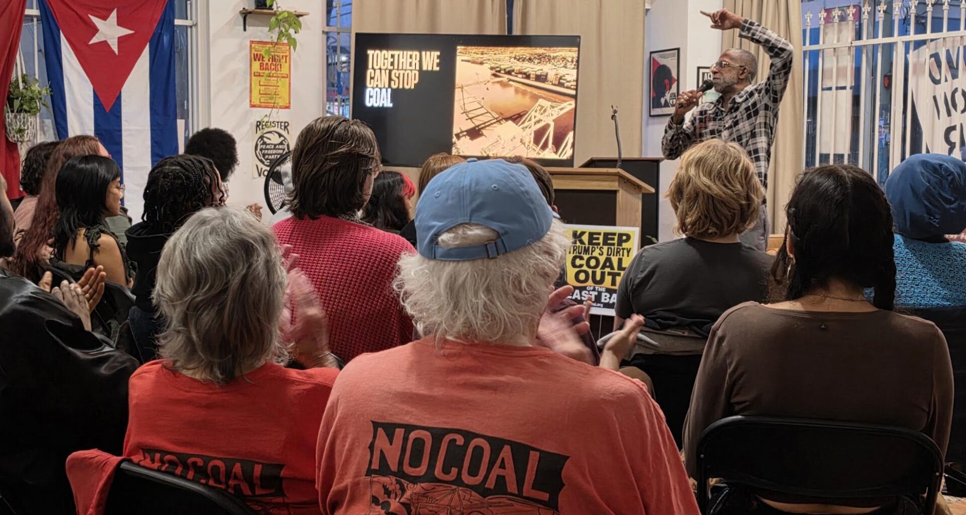 A community organizing meeting with a speaker pointing to a screen with the text "Together We Can Stop Coal in Oakland” as people look on with one attendee wearing a short with the text "No Coal"