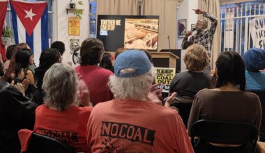 A community organizing meeting with a speaker pointing to a screen with the text "Together We Can Stop Coal in Oakland” as people look on with one attendee wearing a short with the text "No Coal"