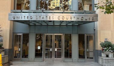 Closeup of the metal canopy over the entrance to a glass-paneled and stone building, with two glass and metal doors at the front and a diamond-shaped stone in a circle on the concrete entryway.