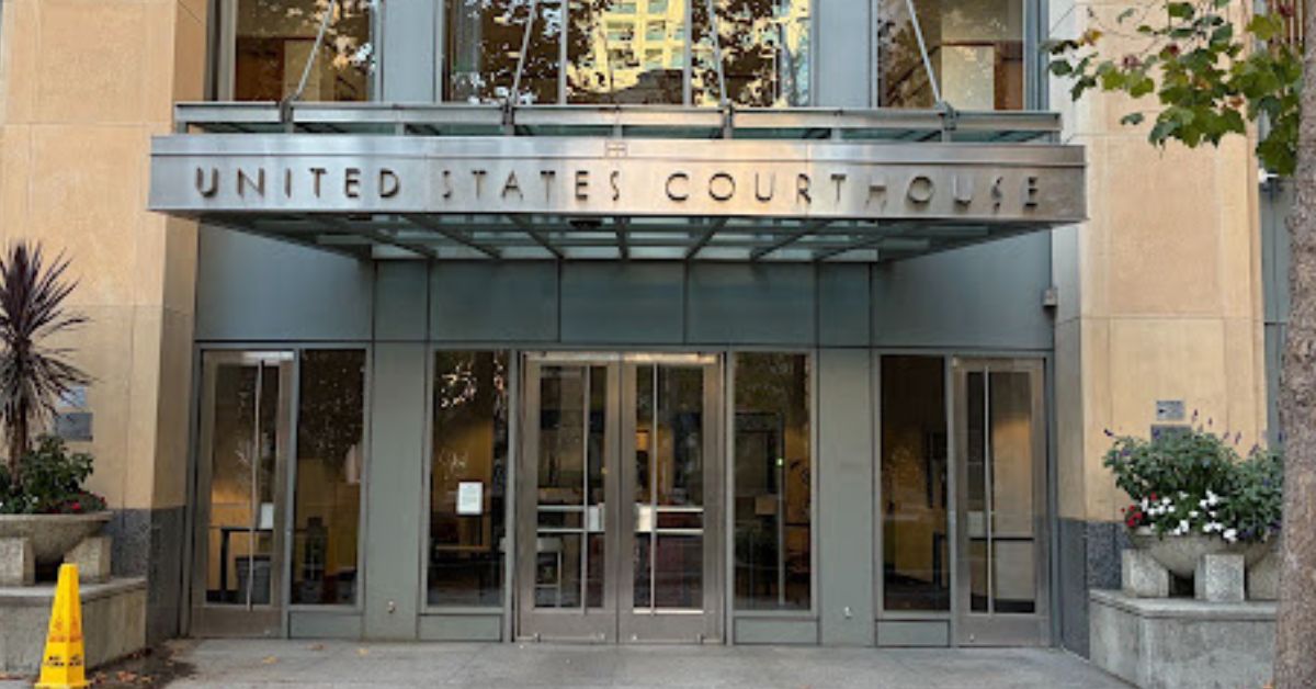 Closeup of the metal canopy over the entrance to a glass-paneled and stone building, with two glass and metal doors at the front and a diamond-shaped stone in a circle on the concrete entryway.