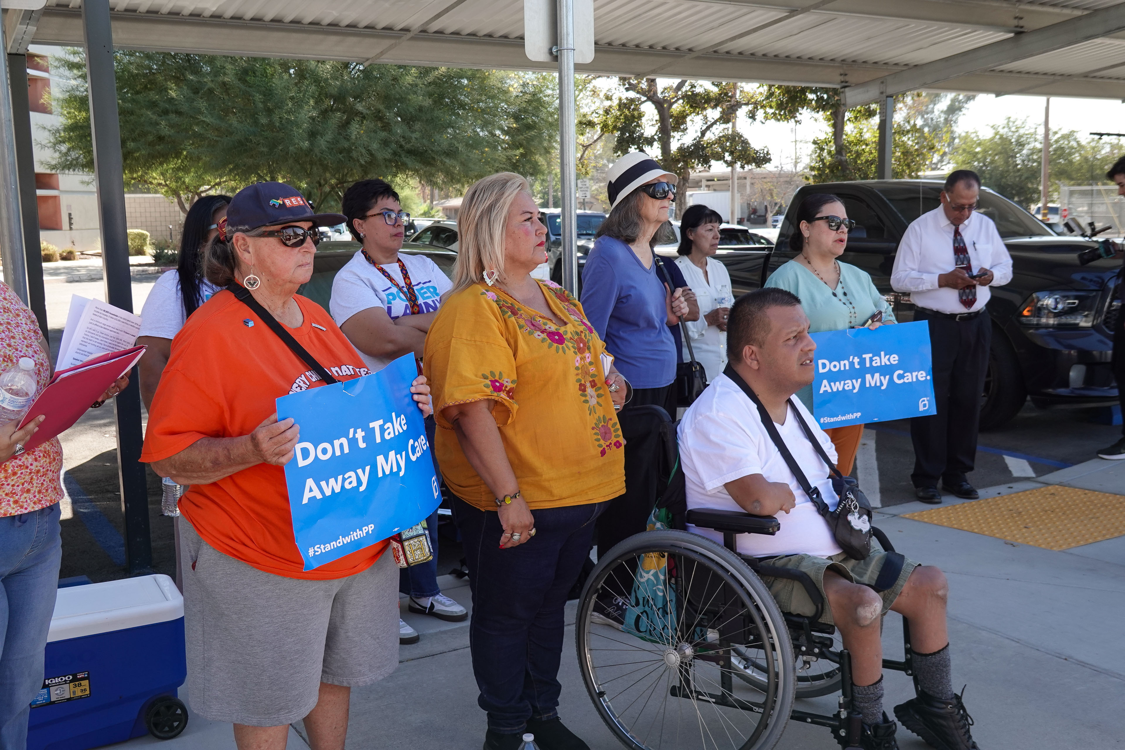 A group of about 10 people stand on a sidewalk. Some hold signs that say, "Don't Take Away My Care. #StandwithPP"