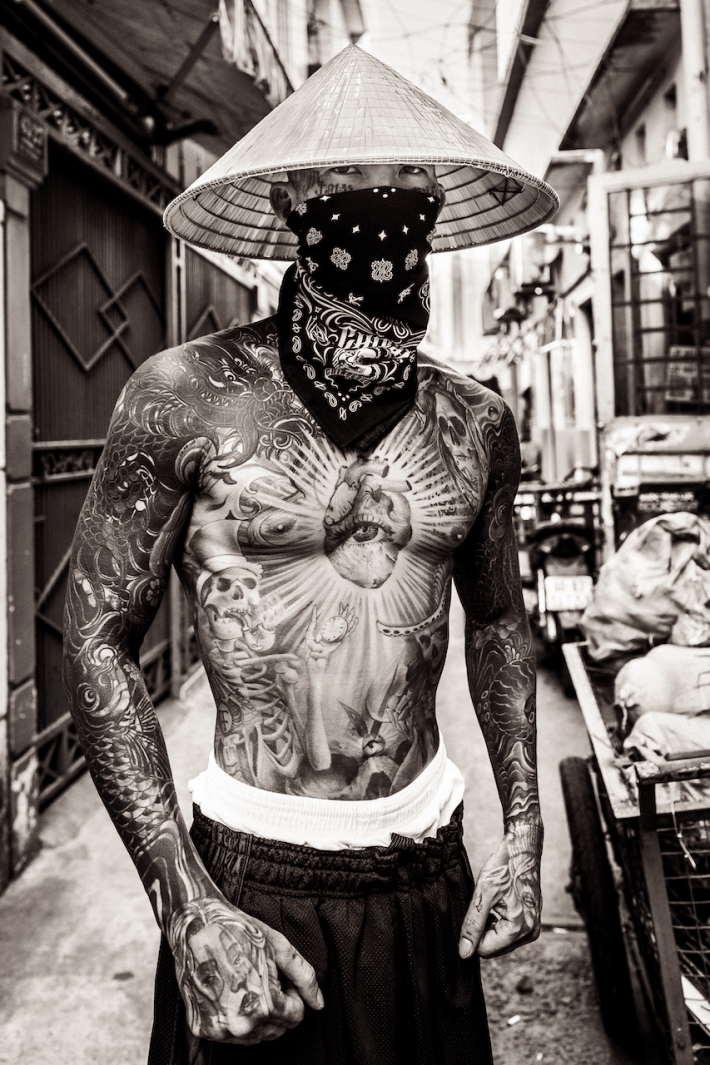 a tattooed shirtless man wearing a conical hat in a street alley
