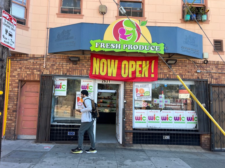 A person with a backpack walks toward the open entrance of a “Fresh Produce” market with “Now Open!” signage and WIC program decals in the windows.