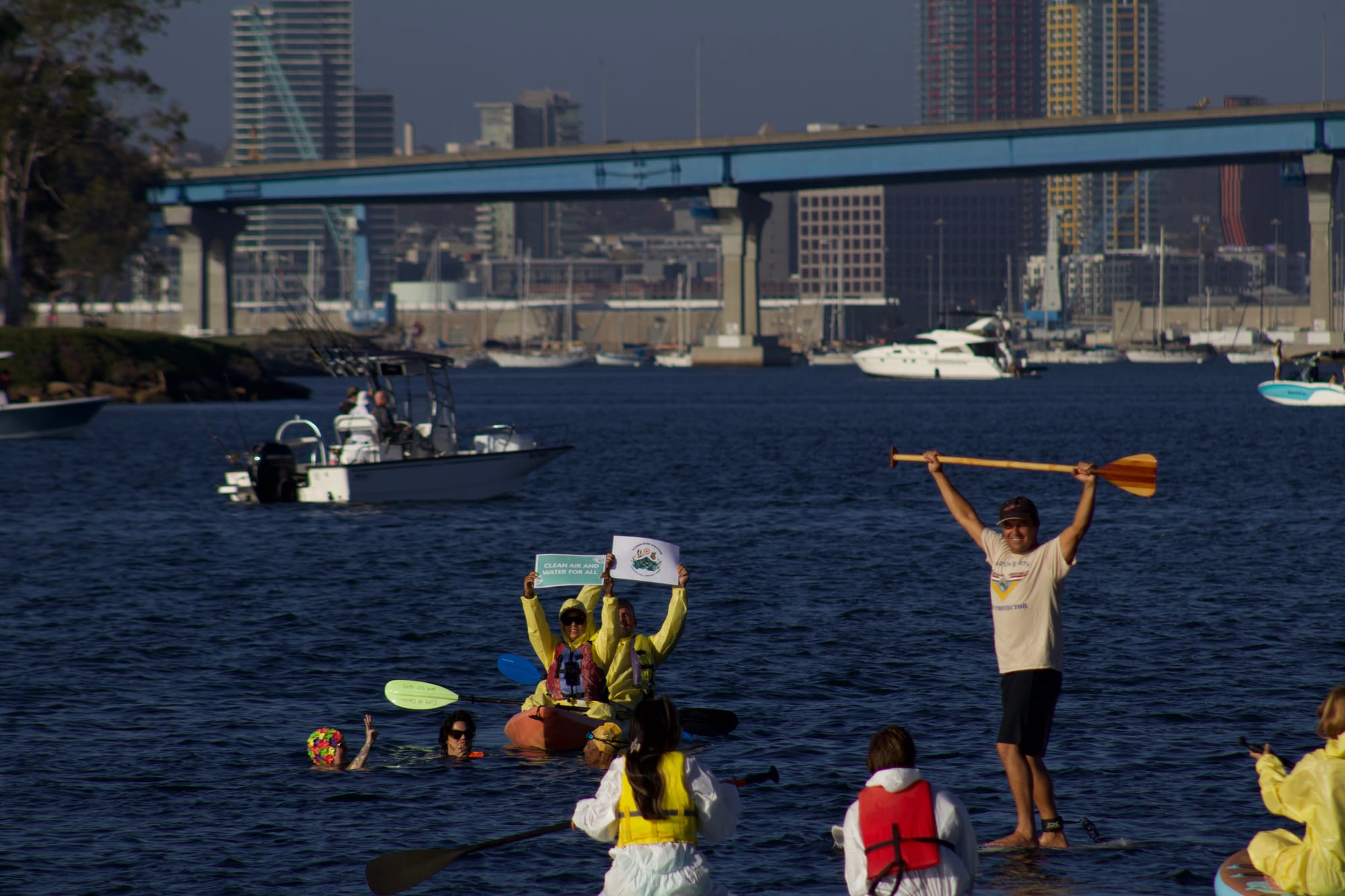 A person on a paddle board holds up a paddle while others sitting down hold signs in front of the Coronado Bridge