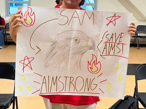 Hands old a hand-drawn sign with a bird in the center and flames and stars around it, with the student's name and "Save AIMS"