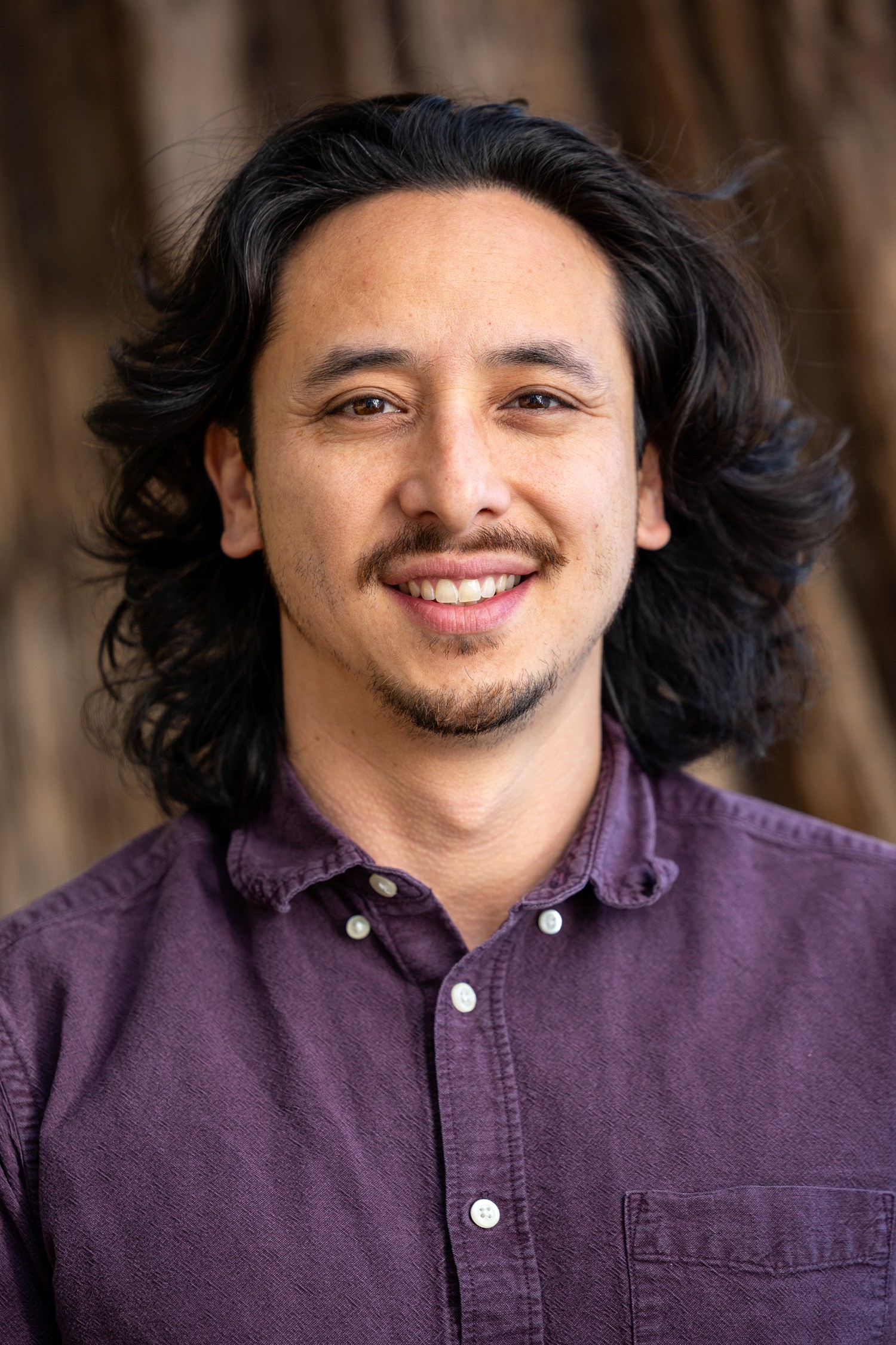 Lucas Zucker's headshot. He is a Latino man with a warm smile, mustache and goatee, and dark flowing shoulder length hair. 