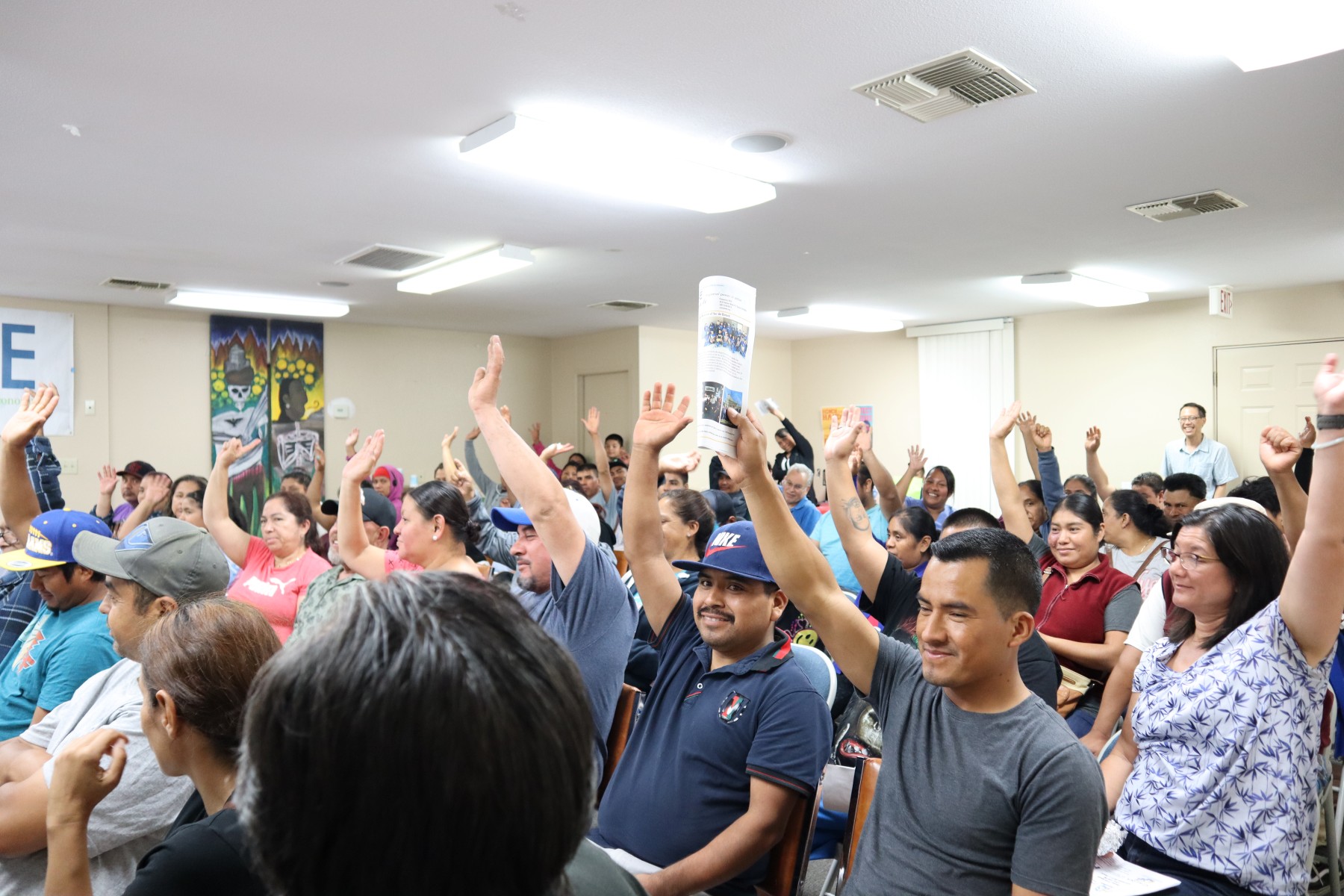 Tens of people seated in rows of seat during a community meeting raise their hands in agreement