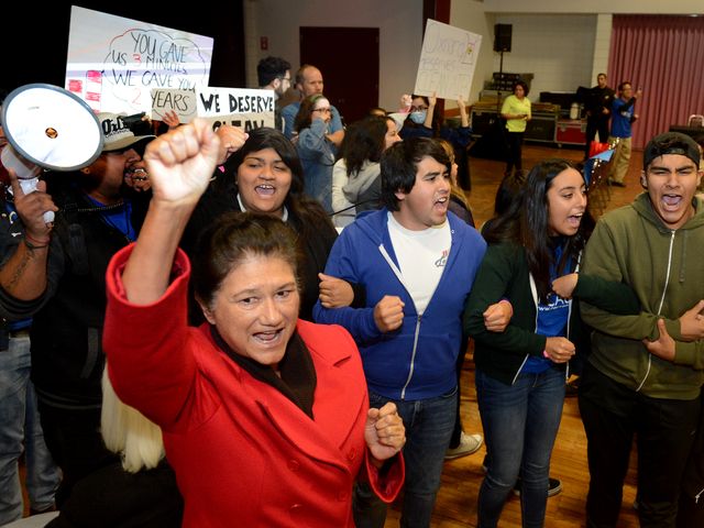 A line of youth activists link arms and chant. In front in sharp focus, an older woman raises her fist powerfully.