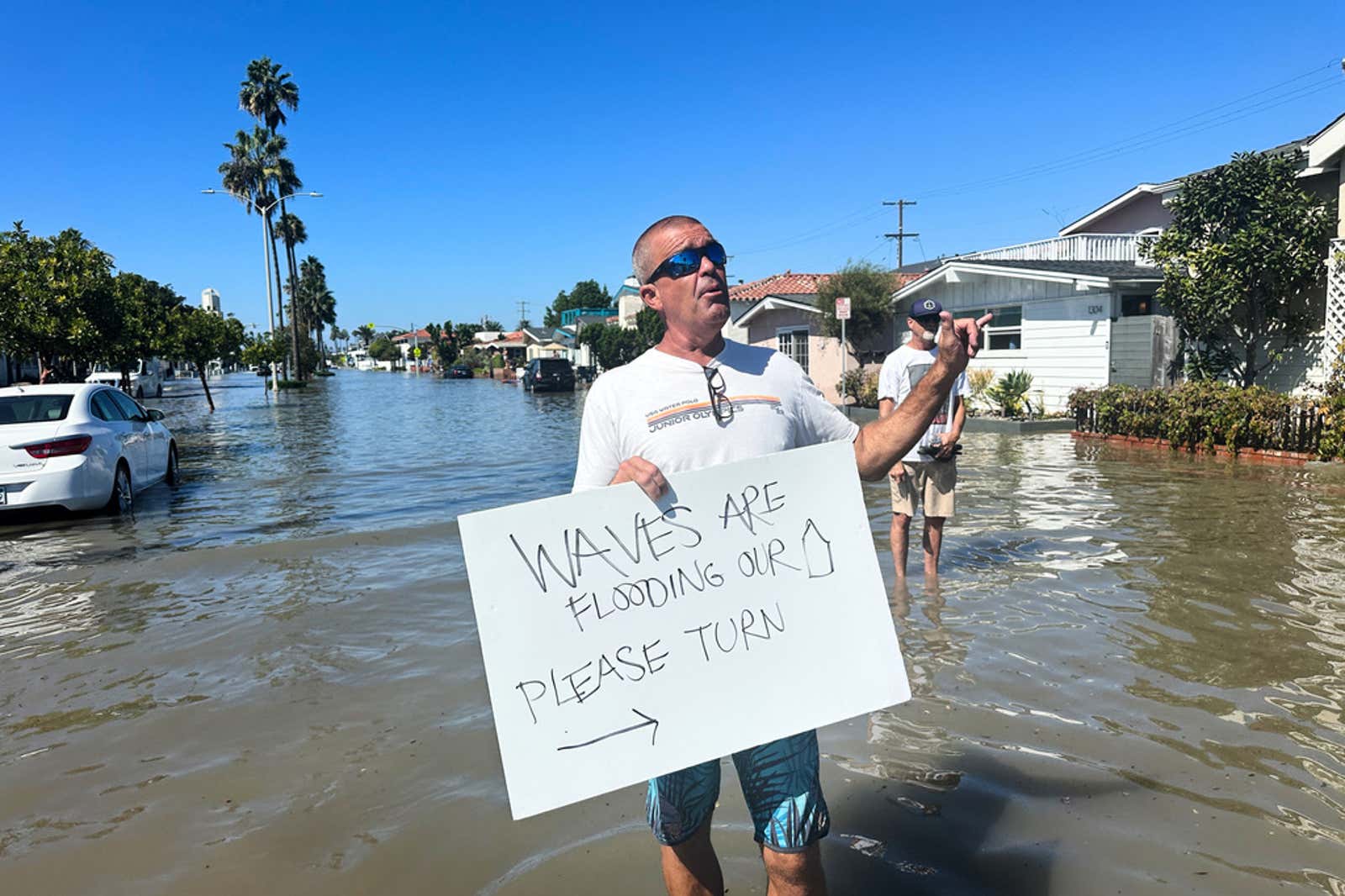 Man holding a sign reading "Waves are flooding our street, please turn" while standing in a flooded residential street with submerged cars and palm trees