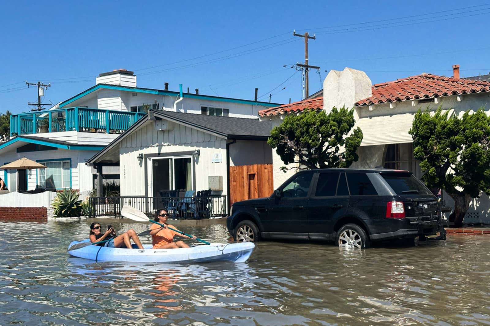 Two people kayaking past a partially submerged black SUV on a flooded residential street