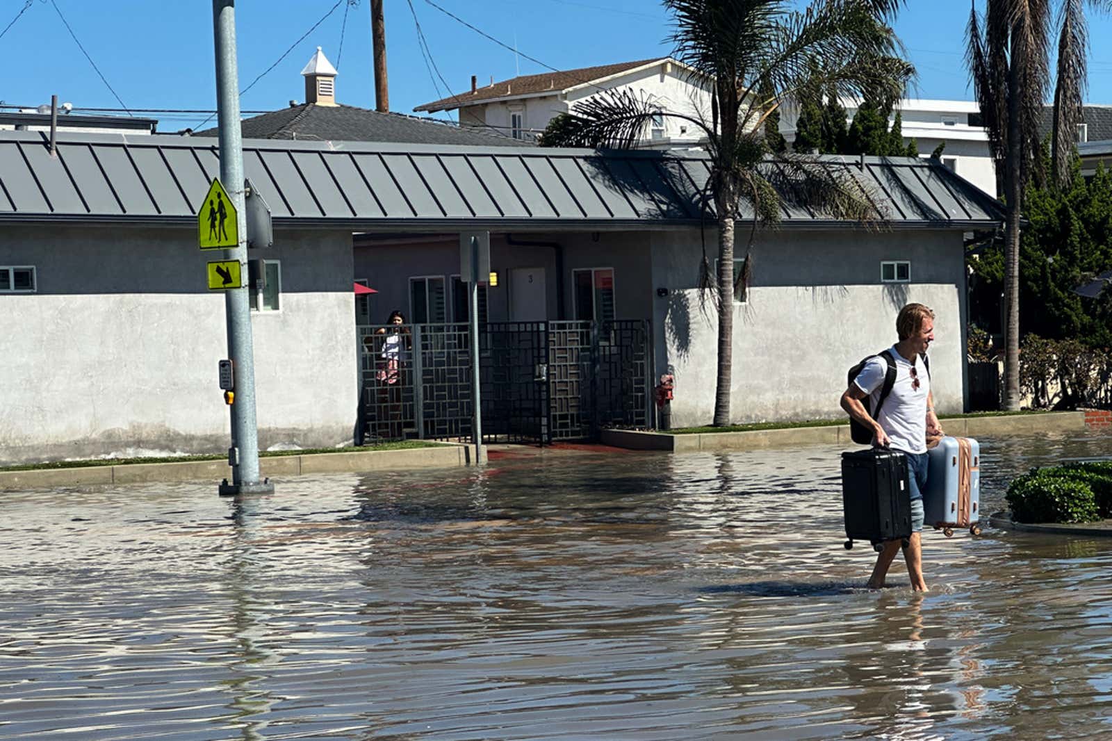 Man wades barefoot through flooded street carrying two suitcases near a gray house with palm trees