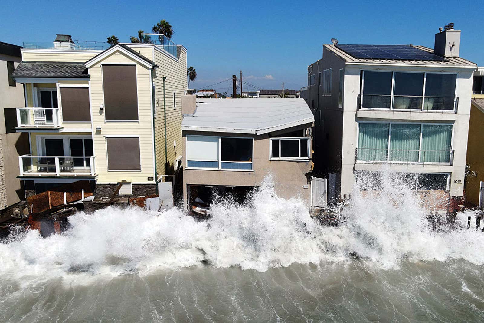Large ocean waves crash against beachfront houses, splashing water onto the buildings' lower floors