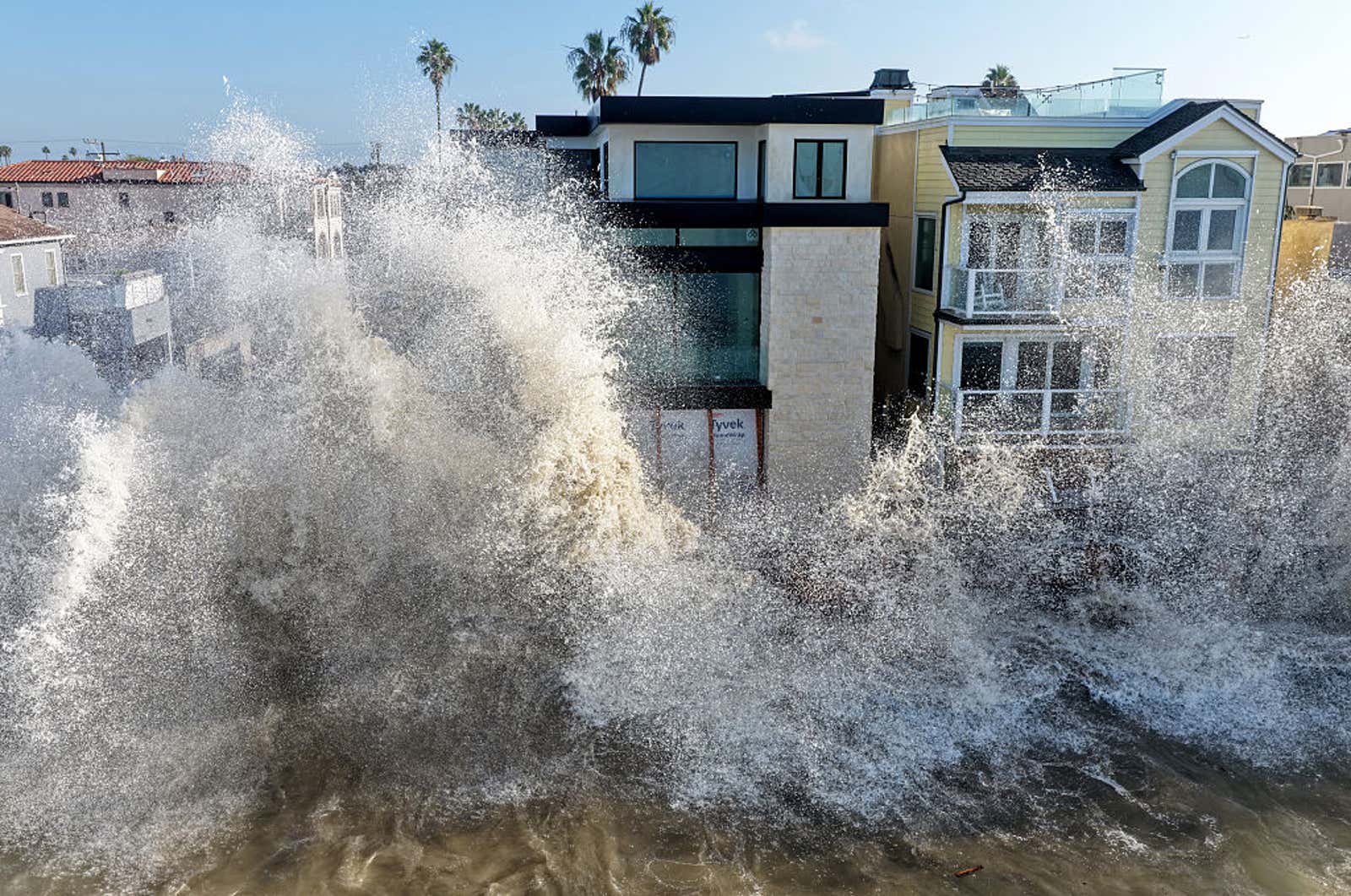 Large ocean wave crashes against beachfront houses, spraying high above the rooftops