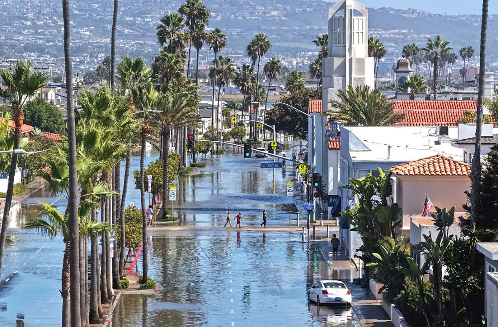 Flooded palm-lined street with submerged road, car partially underwater, and pedestrians wading through standing floodwater