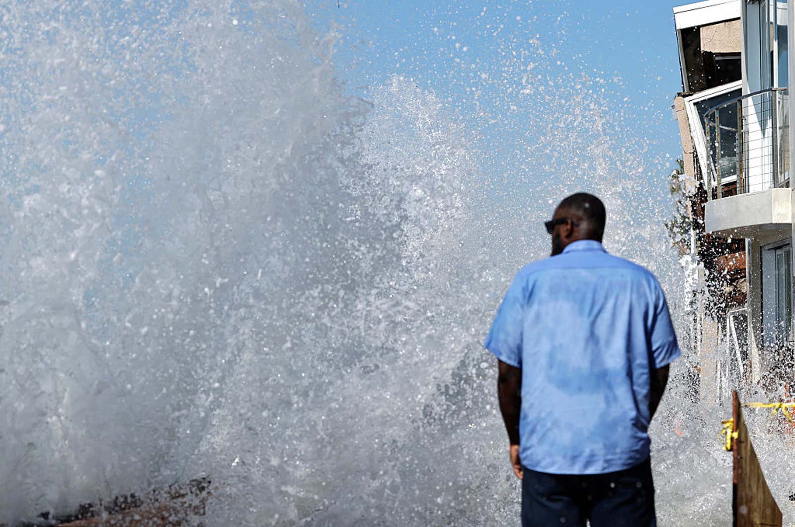 Man in blue shirt watches large ocean wave crash against seawall near coastal buildings