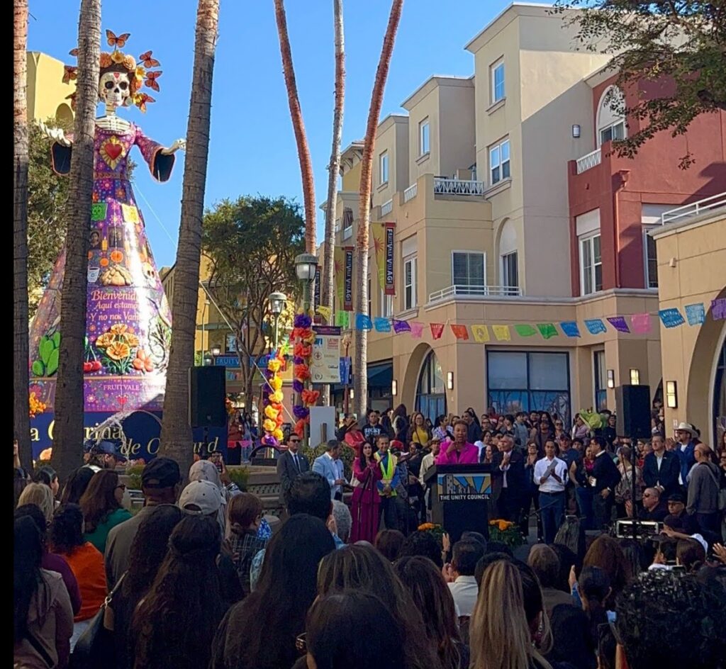 A colossal female skeleton wearing a colorful round skirt and a hate with butterflies protruding towers over a crowd gathered on a city street under pastel-colored banners.