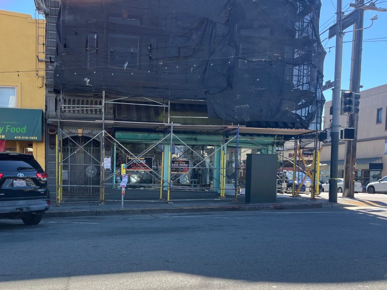 A building under construction with scaffolding and black tarps covers the front. Cars are parked nearby, and a traffic signal is visible at the street corner.