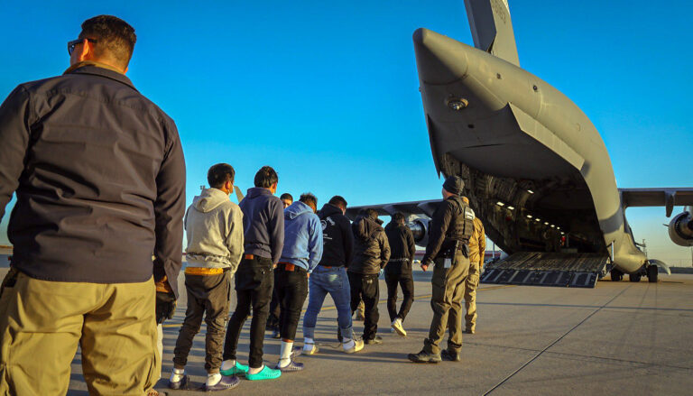 A line of men in chains, seen from behind, are marched by law enforcement officers toward the open back hatch of a massive transport aircraft.