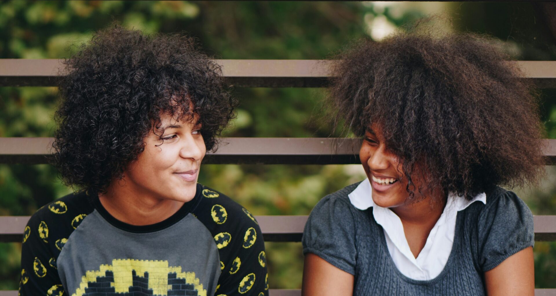 Two Black teens lean against a metal fence and smile at each other