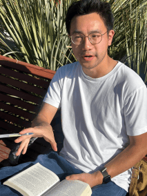 A man in a white t-shirt and glasses sitting on a bench, holding an open book and a pen while gesturing.