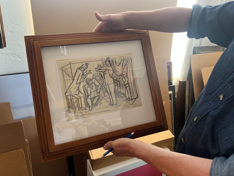 A person holds a framed ink drawing of figures in a theatrical or interior setting.