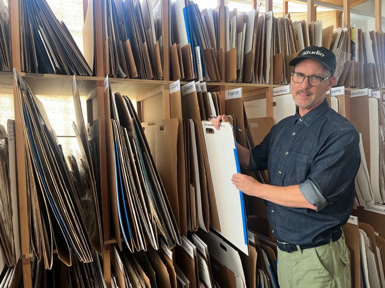 A man in a studio cap and glasses holds a white project board while standing in front of large storage shelves filled with cardboard folders.
