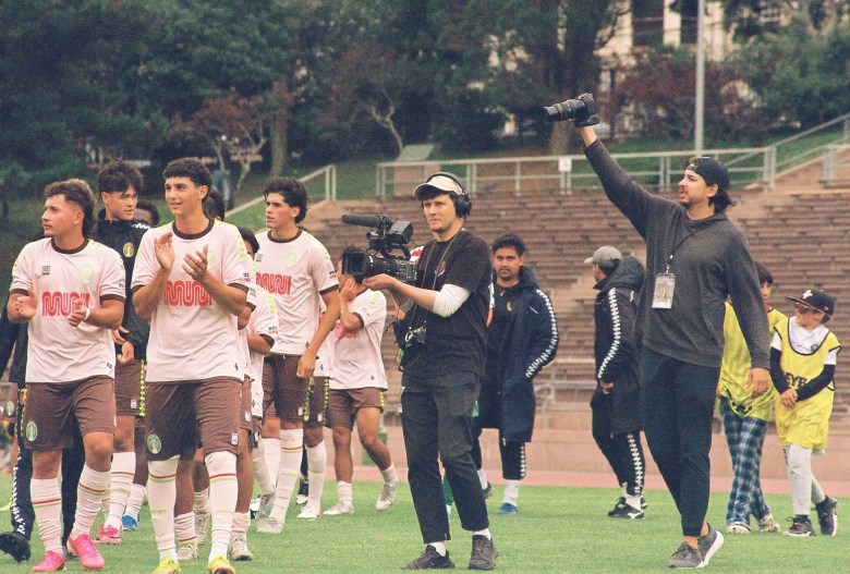 A group of soccer players walks on a field, followed by two camera operators filming them; some people are in the background near stadium seats.