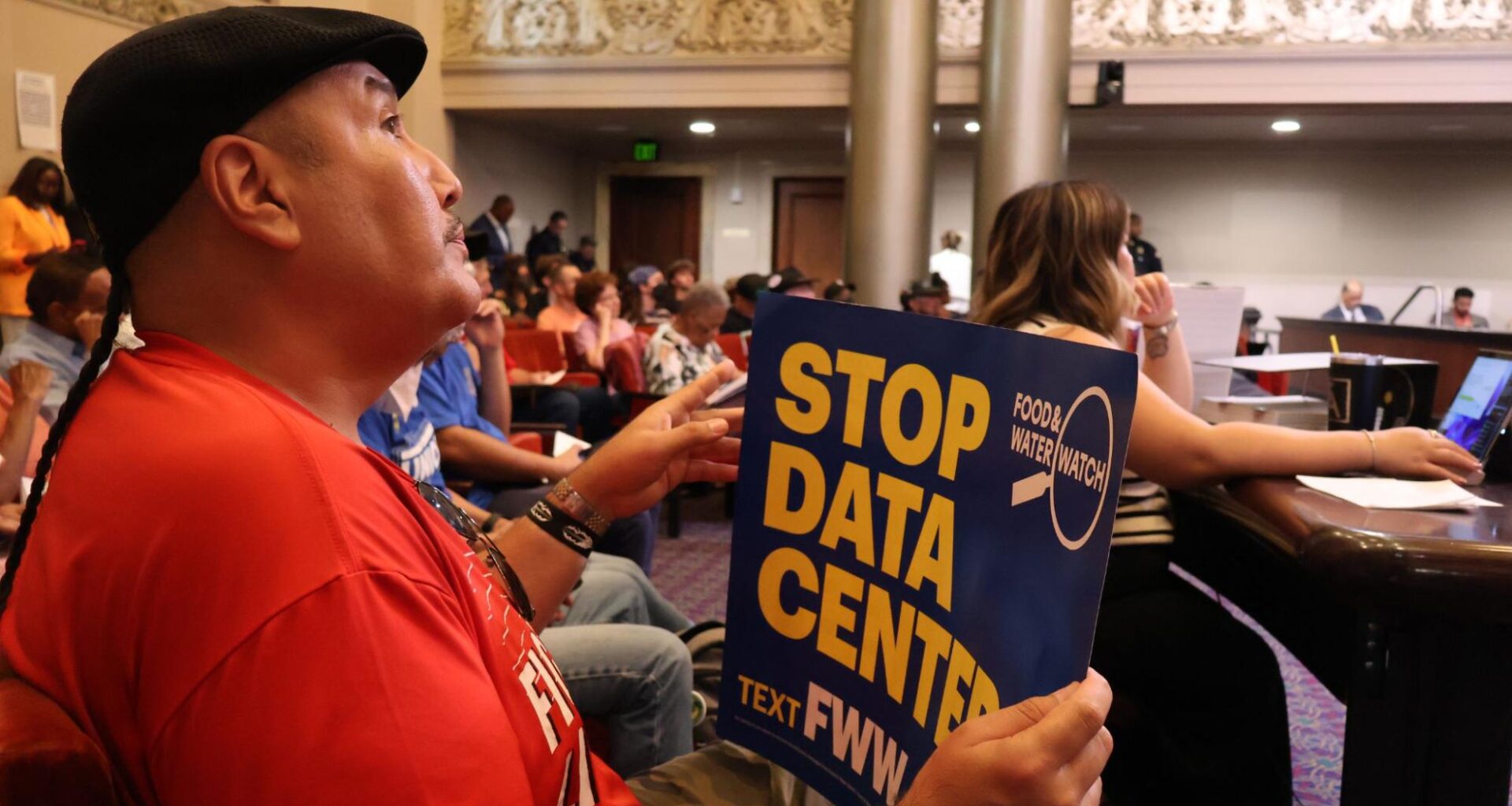 Seated in the audience section of Oakland City Council Chambers, a man wearing a black flat cap and red shirt holds up a blue poster sign reading "STOP DATA CENTER" with the Food & Water Watch logo visible.