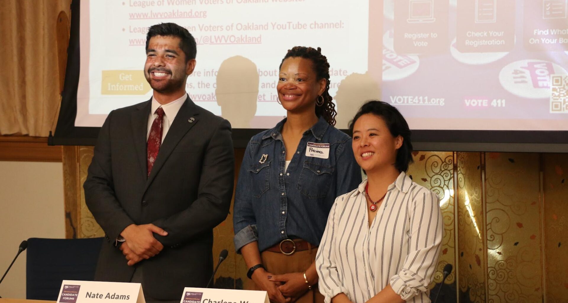 Oakland City Council District 2 candidates stand side-by-side behind a wooden desk following a candidate forum. From left to right: Nate Adams in a dark suit and red tie, moderator Precious Green in a denim button-down shirt with a name tag, and Charlene Wang in a striped button-down shirt on the right.