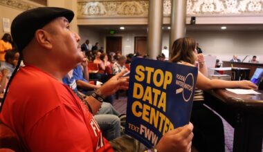 Seated in the audience section of Oakland City Council Chambers, a man wearing a black flat cap and red shirt holds up a blue poster sign reading "STOP DATA CENTER" with the Food & Water Watch logo visible.