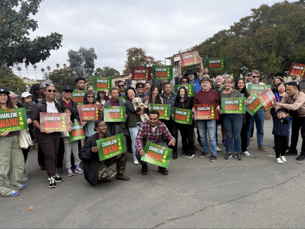 About 30 people with signs line up outside on a pavement smiling at the camera.