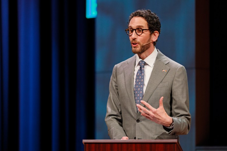 A man in a gray suit and glasses speaks at a podium on stage, wearing a headset microphone, with a blue curtain and a blurred background behind him.