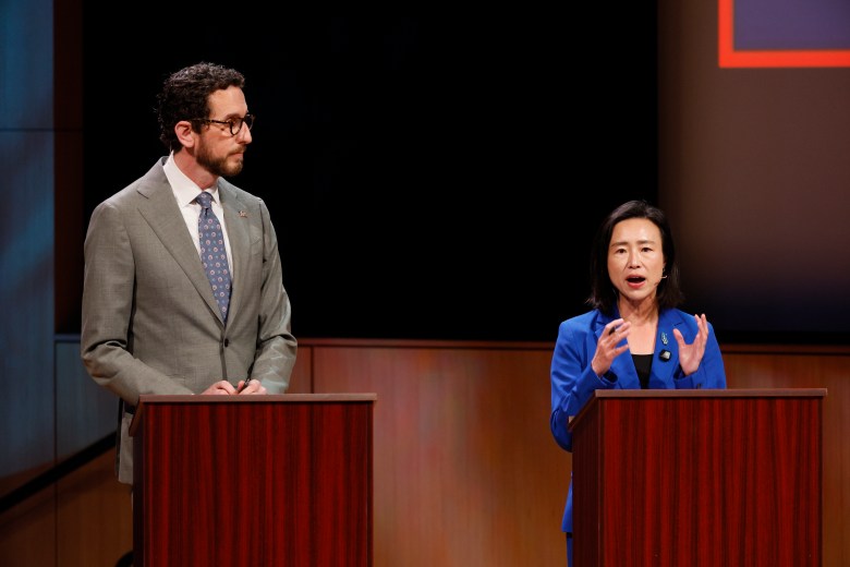 Two people stand behind podiums. The man on the left listens while the woman on the right speaks, gesturing with her hands. Both are dressed in business attire on a stage.