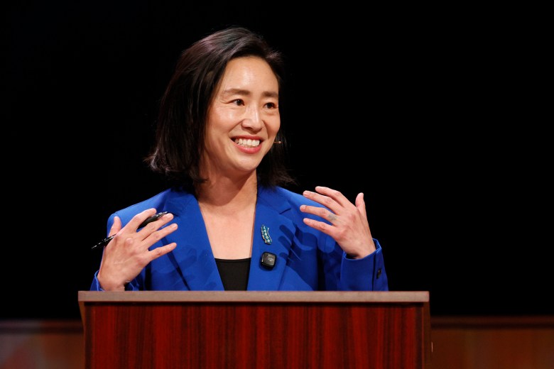 A woman in a blue blazer stands behind a wooden podium, smiling and gesturing with both hands during a speech.