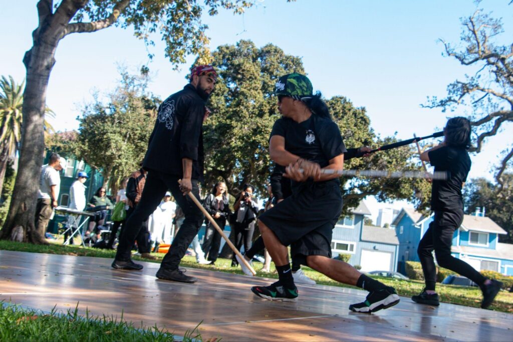 Two men in caps, dressed in black are swinging sticks in motion on a wooden floor on top of grass on a residential street.