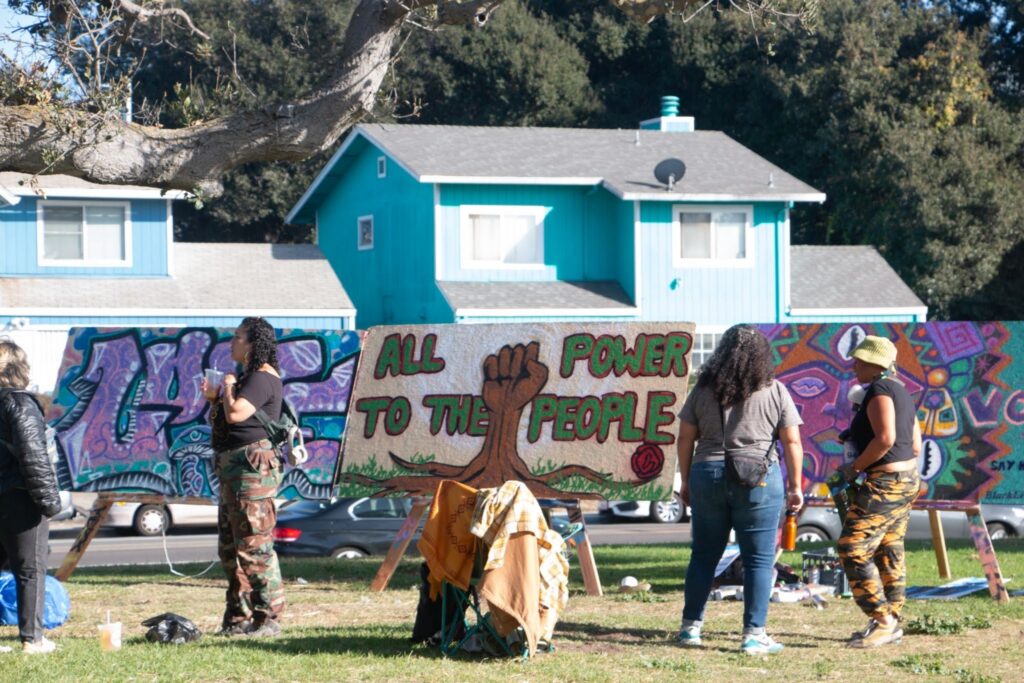 On a patch of grass on a residential street, four people are chatting near a sign that shows a raised brown fist with the words "all power to the people."