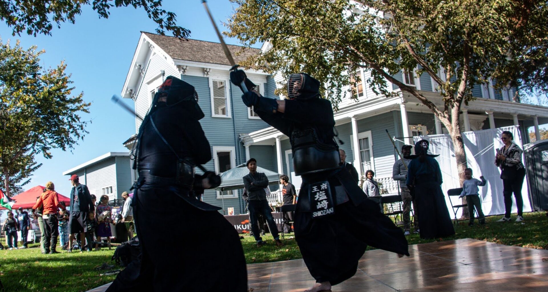Two sworded men in black genie pants and bare feet swing the swords at each other as a sparse crowd looks on outside a house. The men are on a makeshift wooden floor set up on grass.