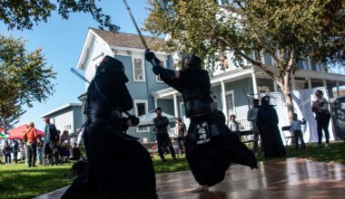 Two sworded men in black genie pants and bare feet swing the swords at each other as a sparse crowd looks on outside a house. The men are on a makeshift wooden floor set up on grass.