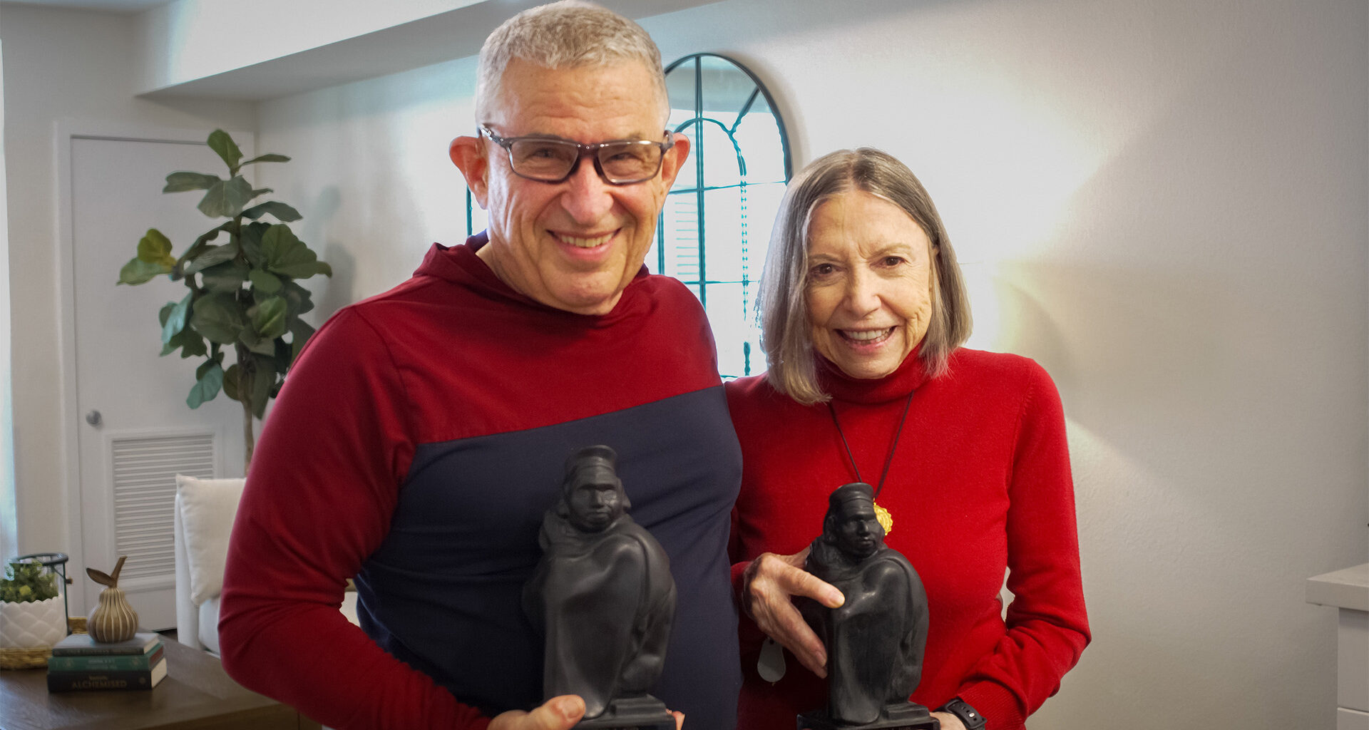A senior couple, both wearing red tops and holding statuettes