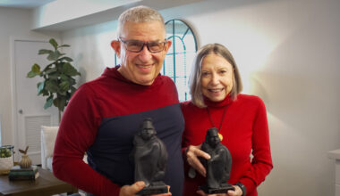 A senior couple, both wearing red tops and holding statuettes