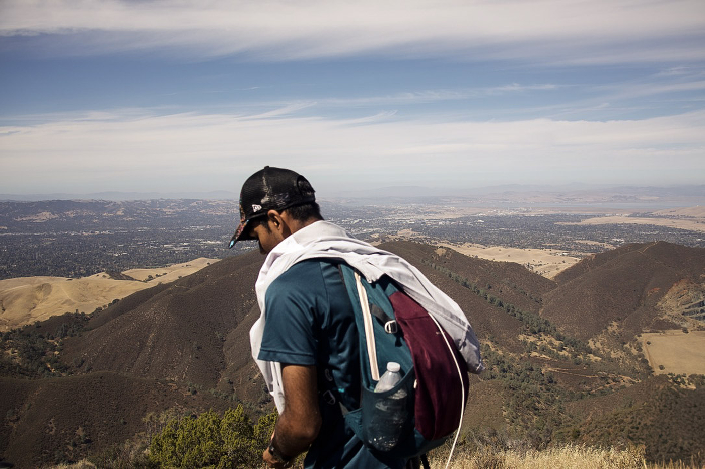 A hiker in a cap and backpack, with a white cloth over the shoulders, looking out over brown hills toward a distant valley.