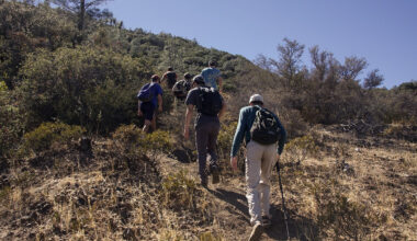 A group of hikers with backpacks climbing a dirt trail up a dry, brushy hillside under a clear sky, one using a trekking pole.