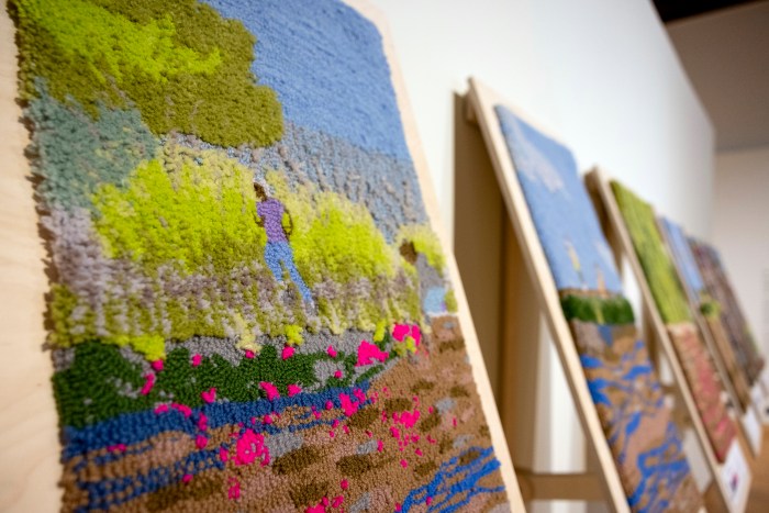 Textile panels with colorful tufted landscapes on angled wooden boards line a gallery wall at the Mills College Art Museum