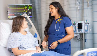 Nurse in blue scrubs smiles while speaking with a patient in a hospital bed with monitors in the background.