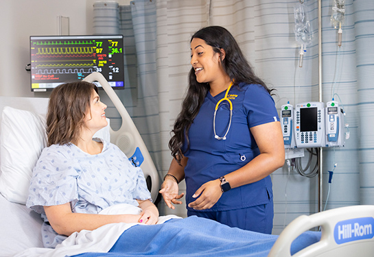 Nurse in blue scrubs smiles while speaking with a patient in a hospital bed with monitors in the background.