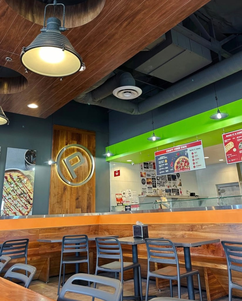 Interior of a shuttered Pieology restaurant, featuring wooden paneling, a large circular "P" logo, and dark tables and chairs.