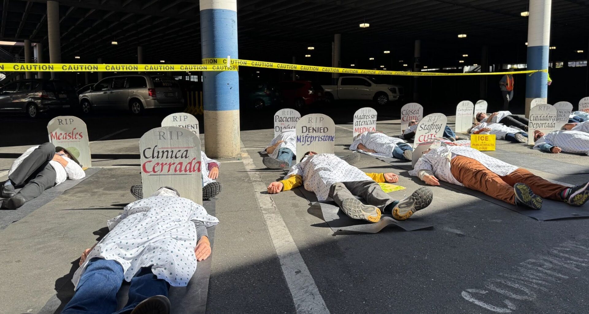 Abot a dozen people are lying in a parking lot in hospital scrubs pretending to be dead. At each one's head there is a tombstone with a name on it. Behind them is police caution tape attached to the parking lot's poles.