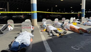 Abot a dozen people are lying in a parking lot in hospital scrubs pretending to be dead. At each one's head there is a tombstone with a name on it. Behind them is police caution tape attached to the parking lot's poles.