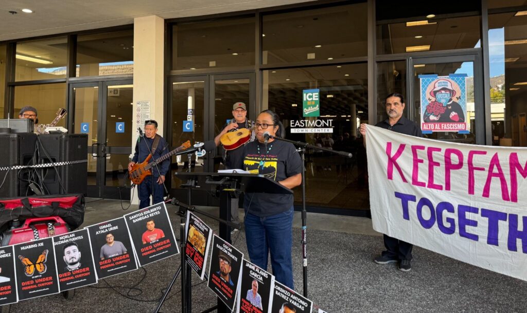 Three guitar players and a man holding a banner stand behind a woman who is at a microphone outside, in front of a glass paneled office buiding. In front of her are posters with people's faces on them.